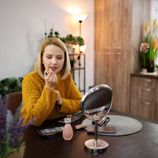 Woman applying makeup in a well-lit room with a mirror and makeup products on a table.